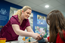 A woman wearing medical clothing measures blood pressure by placing a cuff on the upper arm of a young woman seated at a table.