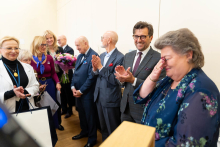 A group of people are standing in a hall during a ceremony; some of them are holding flowers, while others are applauding.