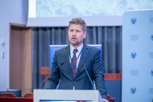A speaker standing behind a lectern with two microphones, wearing a conference badge, addressing the audience, with institutional banners and a projection screen in the background.