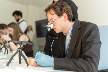 A participant wearing magnifying medical loupes and protective gloves performs a precise procedure on a training model placed on a table, with a small tripod-mounted camera nearby.