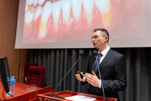 A person standing at a lectern, holding a presentation remote; a slide featuring a large image of teeth is displayed in the background.
