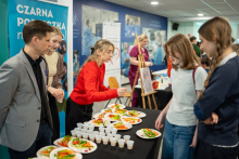 Women and men stand by a table with plates of vegetables and small cups, while one woman serves food portions to event participants.