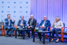 A panel discussion on stage with six seated panelists in armchairs, each holding a handheld microphone or resting hands on their laps, with tables holding water bottles and glasses between the chairs.