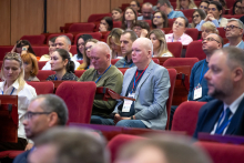 Conference participants seated in red auditorium chairs, some holding notebooks or folders, all facing the stage and listening attentively.