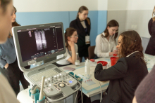 A portable ultrasound machine displays a grayscale scan on its screen while participants sit at a table with medical supplies and containers.
