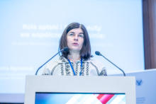 A speaker standing behind a lectern with two microphones, wearing a conference badge, delivering a presentation, with a projected slide containing text in the background.