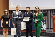 A group stands on a stage during an award ceremony, holding certificates, gift bags, and a presentation case, with a projected event backdrop behind them.