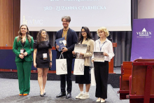 Award recipients stand in a row holding certificates and branded bags on a stage, with sponsor logos and presentation text visible in the background.