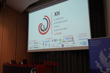 Ekran projekcyjny w sali wykładowej z wyświetloną prezentacją wydarzenia „XII Studenckie Ogólnopolskie Forum Radiologiczne”, wraz z datą 14.03.2026 oraz logotypami sponsorów, partnerów i patronów. Po prawej stronie widoczny roll‑up Warszawskiego Uniwersytetu Medycznego, a na dole scena z mównicą.