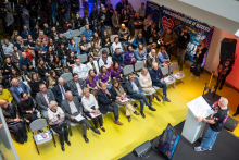 An overhead view of the hall during a charity event. A large audience is seated on chairs facing the lectern, where a speaker is standing. In the background, colorful stage décor can be seen, along with volunteers standing along the walls.