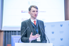 A speaker at a lectern gesturing with one hand while speaking into two microphones, wearing a conference badge, with a presentation slide and institutional logos in the background.