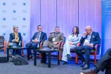 A speaker at a lectern gesturing with one hand while speaking into two microphones, wearing a conference badge, with a presentation slide and institutional logos in the background.