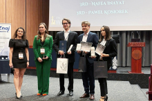 A group of award recipients stands on stage holding certificates and branded gift bags, with a podium and event signage visible nearby.