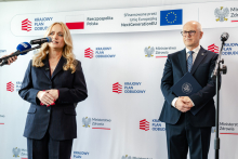 A woman standing at media microphones, dressed in a dark suit, and a man standing next to her, dressed in a suit and holding a folder with the coat of arms in his hands. In the background there is a backdrop with the logos “National Recovery Plan”, “Ministry of Health”, and the European Union.