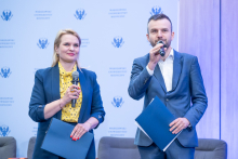Two conference hosts standing on stage in front of a branded backdrop, each holding a handheld microphone and a dark blue folder.