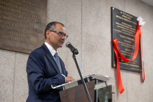 A person in a dark suit stands at a lectern and speaks into a microphone, with a commemorative plaque with red ribbons visible on the wall behind.