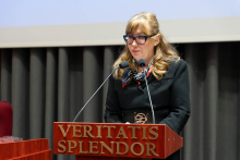 A person standing at a lectern labeled “Veritatis Splendor,” speaking into microphones mounted on the podium; the university emblem is visible on the lectern.