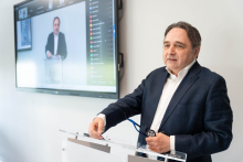 A person standing at a transparent lectern holds a pair of glasses in one hand and rests the other hand on the lectern top. In the background, a screen displays a live feed of the event, including a view of the person speaking.