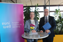 A woman and a man stand at a small round table indoors. The woman holds a bouquet of flowers, and the man holds a blue folder. On the table there are documents and pens. Next to them there is a vertical banner with the logo “European University for Well-Being,” and behind them there are large windows and green plants.