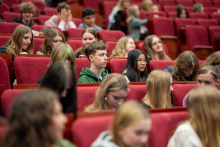 Young people sit in red auditorium seats facing the stage; some of them hold notebooks or mobile phones.