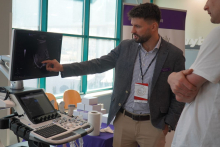 Stanowisko demonstracyjne z aparatem USG: osoba wskazuje na obraz ultrasonograficzny wyświetlany na monitorze, omawiając szczegóły badania. Obok widoczna jest konsola aparatu oraz elementy stoiska edukacyjnego w jasnej sali.