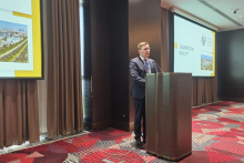 Osoba stoi przy mównicy i wygłasza wystąpienie, trzymając dłonie na pulpicie. Obok mównicy znajduje się ekran prezentacyjny z tekstem „Shaping the Future” oraz grafiką miasta. Sala konferencyjna ma zasłonięte zasłony i wzorzystą wykładzinę.