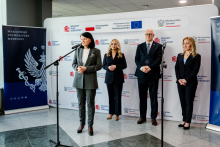 A woman standing at media microphones, dressed in a dark suit, and a man standing next to her, dressed in a suit and holding a folder with the coat of arms in his hands. In the background there is a backdrop with the logos “National Recovery Plan”, “Ministry of Health”, and the European Union.