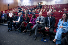The front rows of an auditorium filled with conference participants seated in red chairs, several attendees holding notebooks, folders or pens, with a full audience visible in the background.
