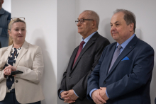 A person in a dark suit stands at a lectern and speaks into a microphone, with a commemorative plaque with red ribbons visible on the wall behind.