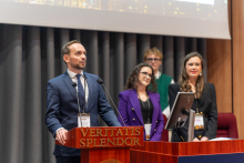 A person speaks at a wooden lectern labeled “Veritatis Splendor,” while several people stand behind, one of them standing next to a computer monitor.