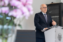 A person stands behind a lectern with the emblem of the Medical University of Warsaw and makes a hand gesture while speaking. In the foreground, on the left side of the frame, there is an out-of-focus bouquet of pink flowers. In the background, a server rack and a light-colored wall of the room are visible.