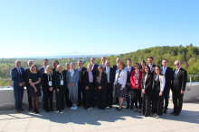A large group of women and men pose for a group photo outdoors on a terrace. The participants are dressed in formal and business-style clothing. In the background there is a landscape with trees, hills, and a clear sky.