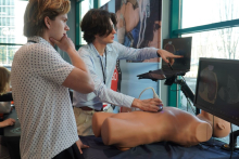 Dwie osoby prezentują badanie ultrasonograficzne na fantomie tułowia; jedna obsługuje głowicę USG, druga wskazuje obraz na monitorach wyświetlających przekroje anatomiczne