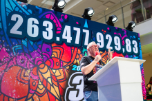 A person standing at a lectern is delivering a speech during a charity event. Behind them, there is a large, colorful screen with dynamic graphics and the displayed amount of 263,477,929.83.