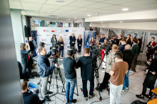 A conference hall with numerous cameras on tripods. In front of a backdrop with logos there are speakers, and in front of them a group of journalists and camera operators recording the event