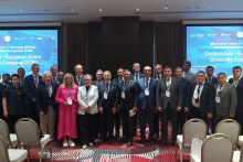 A large group of conference participants standing in rows in a meeting room, wearing formal attire and conference badges, with presentation screens showing the event title behind them.