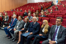 Audience members sit in red auditorium chairs, facing the stage, with some people holding folders on their laps.