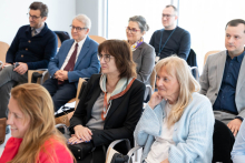 A group of participants sits on chairs arranged in rows in a conference room. The people are facing the lectern, and some have their hands resting on their laps or on the armrests. Large windows allowing daylight into the room are visible in the background.