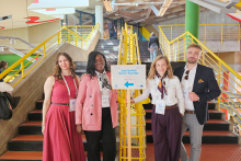 Five adults, three women and two men, stand at the bottom of indoor stairs in a modern building. One woman holds a sign with an arrow and the text “10th EUNIWELL Rectors’ Assembly.” All five wear conference badges on lanyards, and yellow railings and staircases are visible behind them.
