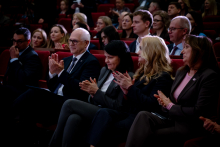 A row of the audience seated in red auditorium chairs. Women and men are applauding, holding their hands raised in front of them. The people are dressed in formal clothing.
