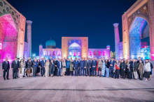 A wide group photo of conference participants standing outdoors at night in front of illuminated historic architecture with arches and domes, arranged in a long line across a stone courtyard.