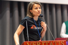 A person speaks into a microphone at a lectern, with one hand resting on the chest and a name badge hanging on a lanyard.