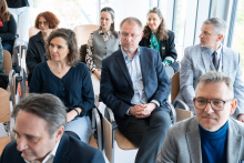 Event participants sit on chairs in a bright conference room. People in the foreground have their hands folded on their laps, while other attendees are visible in the rows behind them. Large windows with a view outside are located on the right side.