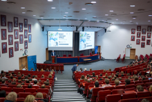  A view of a lecture hall filled with young people seated in red chairs; a lectern and a presentation screen are visible on the stage.