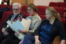 Trzy osoby siedzą obok siebie na czerwonych fotelach w sali audytoryjnej; jedna z nich trzyma bukiet kwiatów oraz dokumenty, co sugeruje udział w uroczystości lub wydarzeniu oficjalnym.