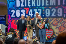 A group of four people pose for a joint photo against the backdrop of a large, colorful screen displaying the word “Thank you!” and the amount 263,477,929. In the foreground, a photographer can be seen taking pictures during the charity event.
