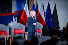 A woman standing on stage at a microphone, dressed in a dark suit. The woman is holding her hands clasped in front of her. In the background there are the flags of Poland and the European Union, as well as armchairs and a table with a bottle of water.