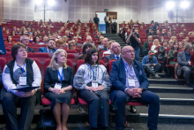 Conference participants seated in red auditorium chairs, listening attentively, some holding notebooks or conference materials, with cameras and photographers positioned in the back rows.