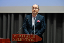 A person standing at a lectern, speaking into two microphones; a sheet of paper lies on the podium, with auditorium curtains visible in the background.