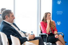 Two people sit on chairs arranged in the event space. One person holds a water bottle, while the other rests a hand on the chin. In the background, there is a blue backdrop with the repeated logo of the Medical University of Warsaw.